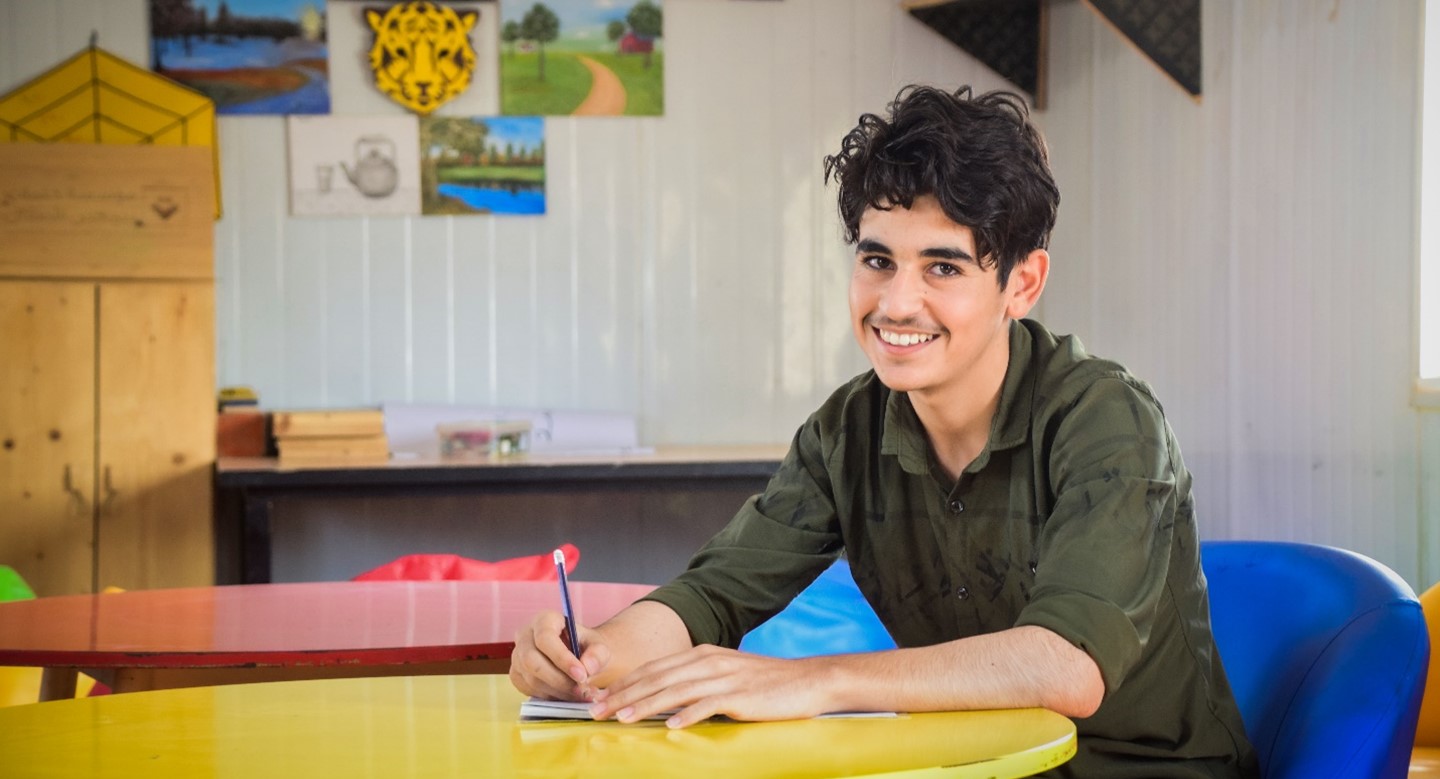 A refugee youth (boy) sitting at a desk holding a pen