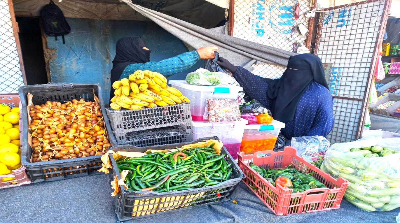 a woman at a vegetable stand hands a bag to another woman