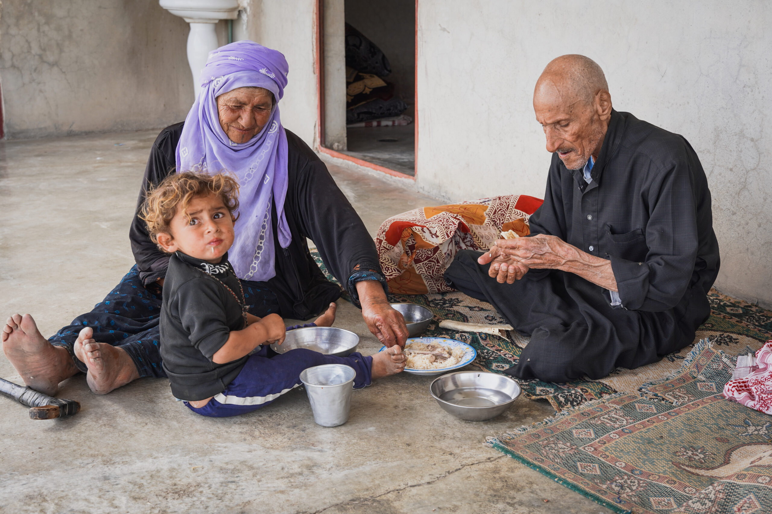 an older woman, older man, and young child sit on the ground eating a meal together