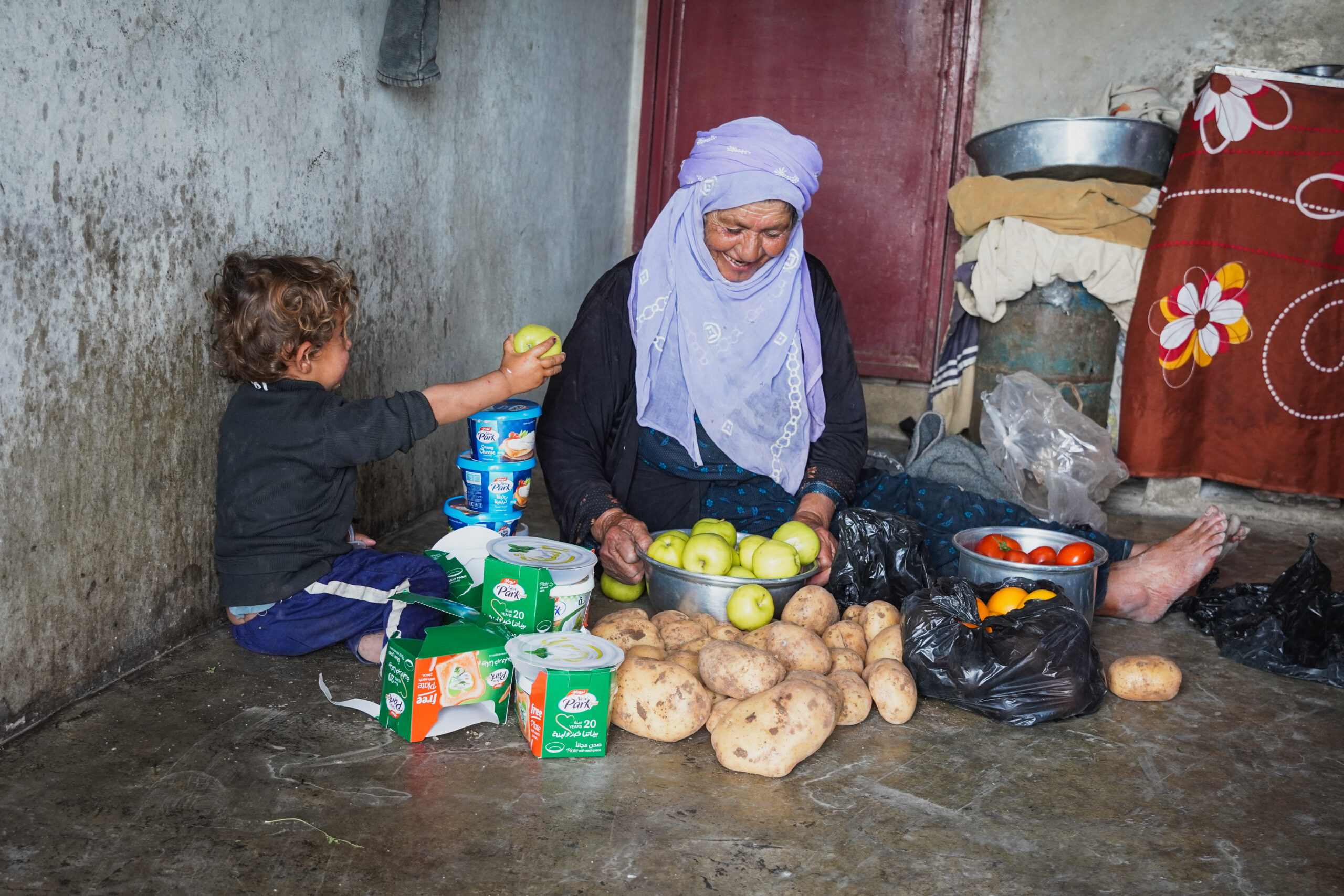 an older woman sorts through fresh produce on the floor of her kitchen alongside her grandson