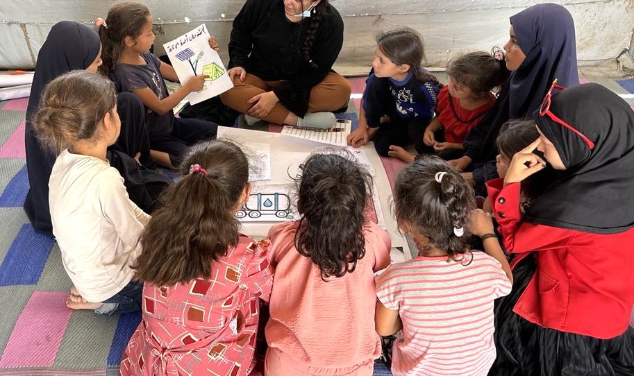 children sit in a circle in a tent looking at a poster